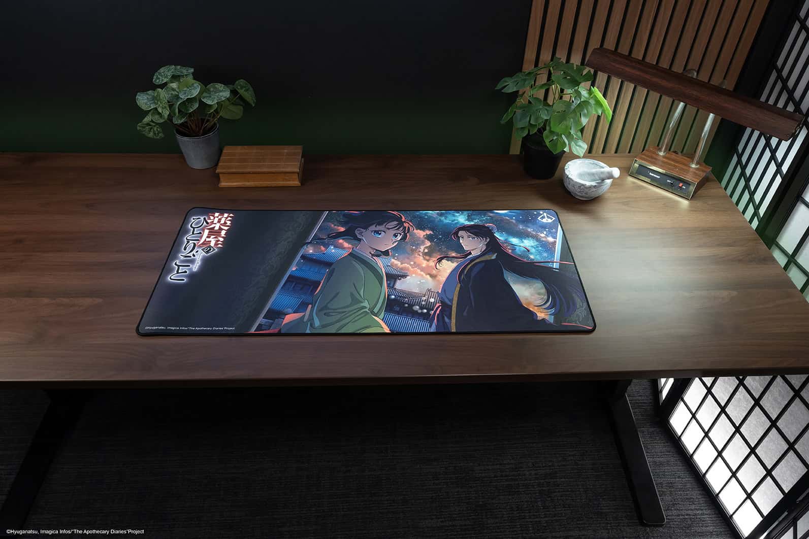 041326-Apothecary_Diaries-social-16_2x | The Outerhaven Long wooden desk with a large anime-themed mouse pad showing two characters against a night sky; potted plant and small wooden boxes sit at the back left, and a lamp and mortar bowl are on the right edge.