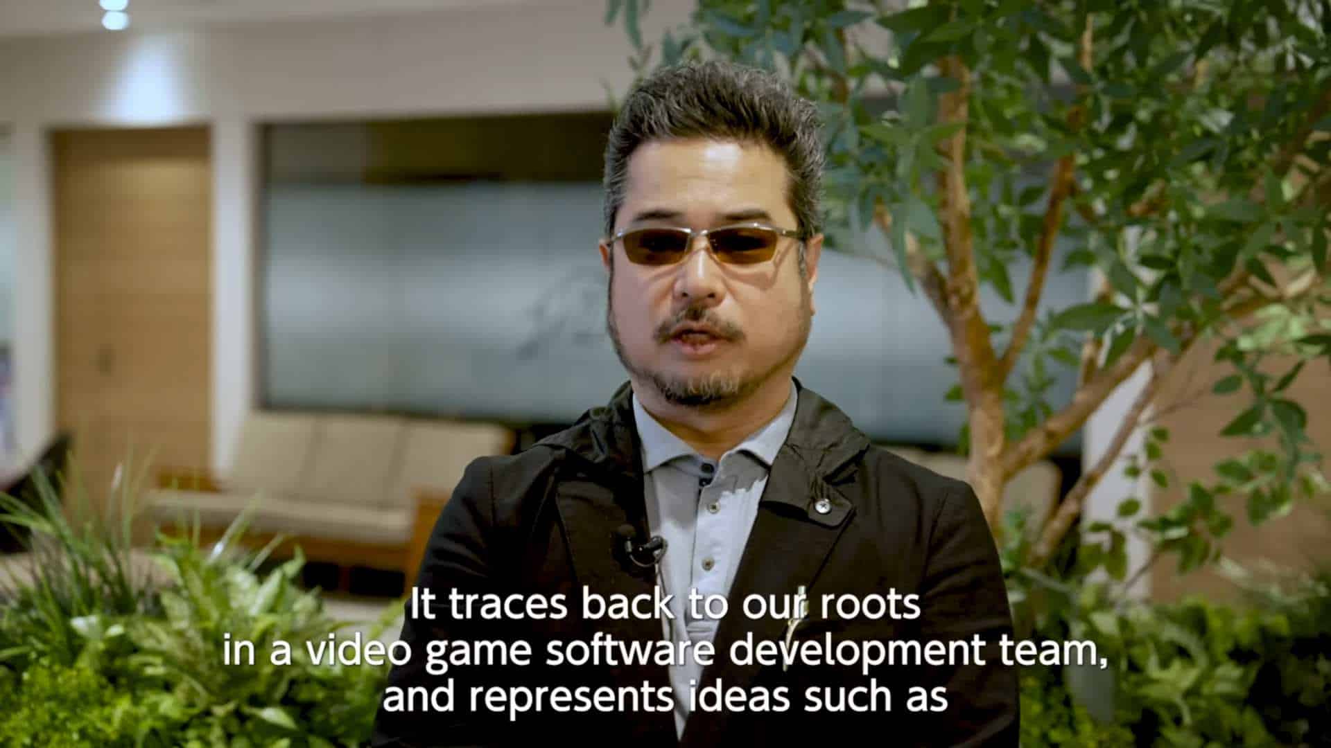 New Studio VS Studio SNK Announced _ A Message from Katsuhiro Harada 1-16 screenshot | The Outerhaven Man wearing sunglasses and a blazer speaks on camera in a plant-filled indoor setting; white subtitles are visible at the bottom.