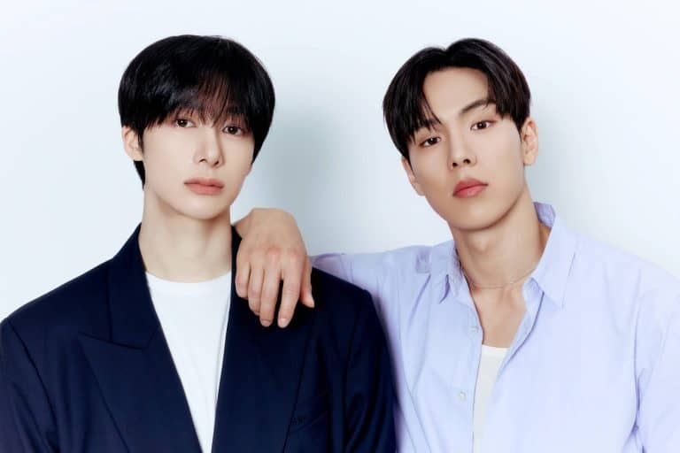 SHOWNU X HYUNGWON_1 | The Outerhaven Two young men pose closely together against a pale background; one in a dark blazer, the other in a light lavender shirt, with a hand resting on the shoulder between them.