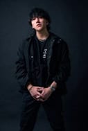 TeddyLoid | The Outerhaven Young man with dark hair wearing a black jacket stands against a dark studio backdrop, hands clasped in front.