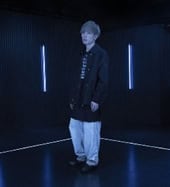 hachi | The Outerhaven Portrait of a person standing in a dark studio with blue vertical light bars behind them, wearing a dark jacket and light pants.