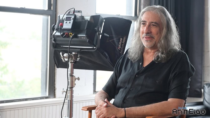 Mike Pascuzzi | The Outerhaven Man with long gray hair in a black shirt sits beside a large studio light on a stand in a bright room.