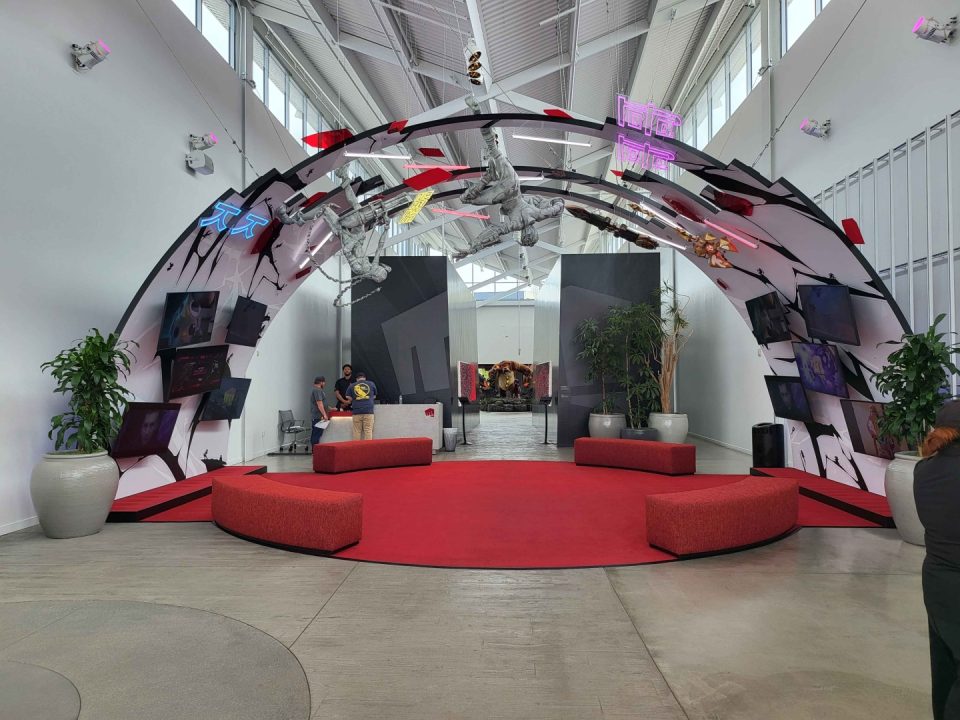 Indoor exhibition lobby with a red circular carpet under a curved archway featuring astronaut figures, neon signs, wall-mounted screens, and potted plants; a reception desk and visitors in the background.