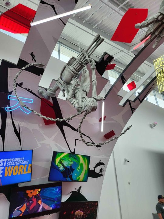 Suspended skeleton figure holding a large gun, chained to the ceiling among neon panels and game-themed wall art in a gallery. Coalition of sci-fi display with screens below.