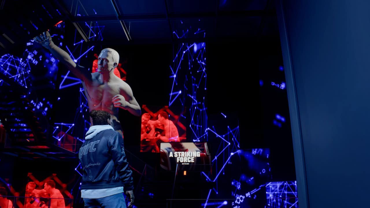 UFC6HoL | The Outerhaven Person in a blue jacket stands before a large digital display of a shirtless statue raising an arm, with blue neon network visuals and a panel reading 'A STRIKING FORCE'.