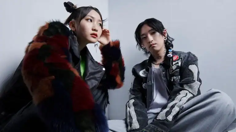 yoasobi-clockenflap | The Outerhaven Two stylish young adults sit against a pale wall: a woman in a multicolor faux fur coat and a green top on the left, a man in a patch-covered denim jacket on the right.