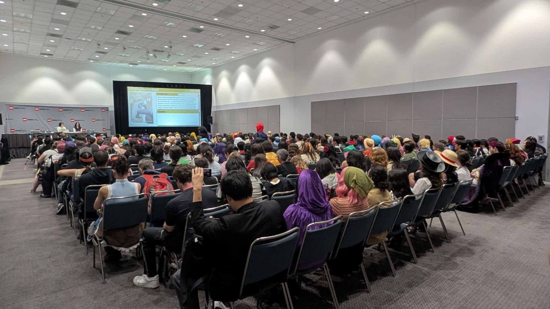 Manga Plaza Anime Expo | The Outerhaven Large conference hall with a stage and screen; a panel of two people sits at a long table while a crowded audience watches a presentation.