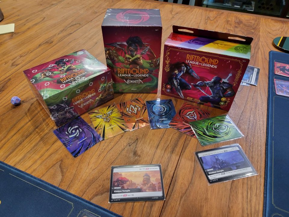 1000019612 | The Outerhaven Colorful Riot Bound: League of Legends trading card game boxes and cards spread on a wooden table, with a blue die nearby.