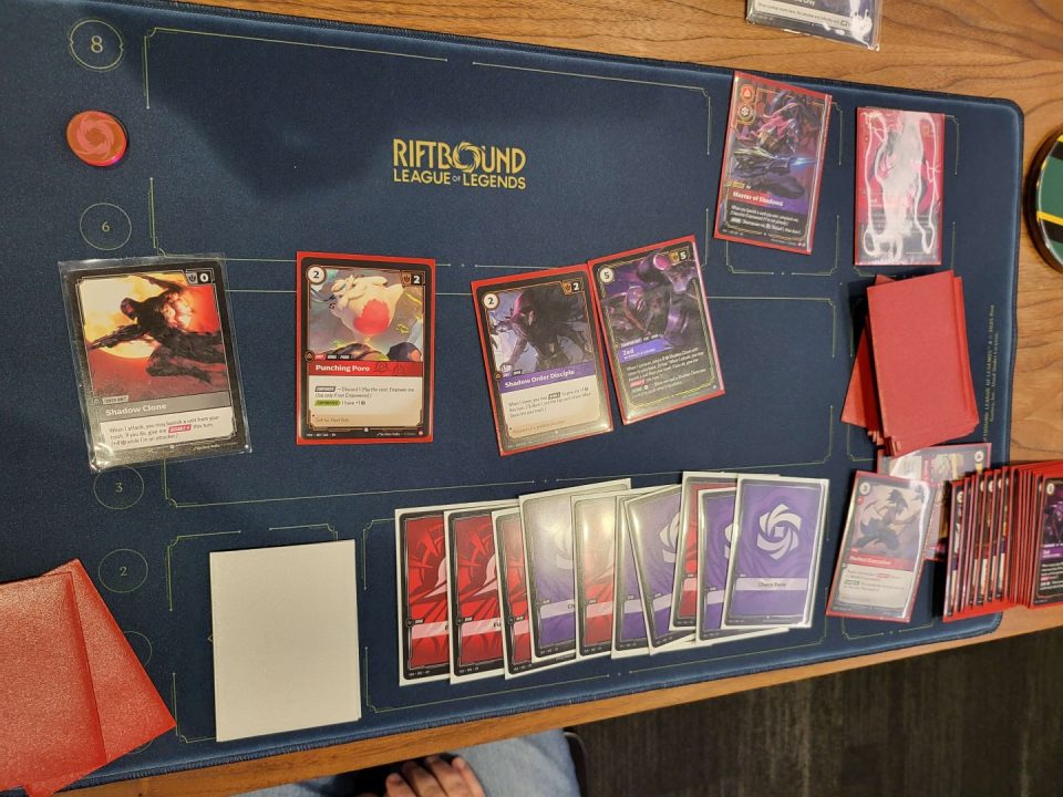 1000019615 | The Outerhaven A Riftbound League of Legends card game table with red-bordered cards laid out in a row and a stack of red card sleeves to the right.