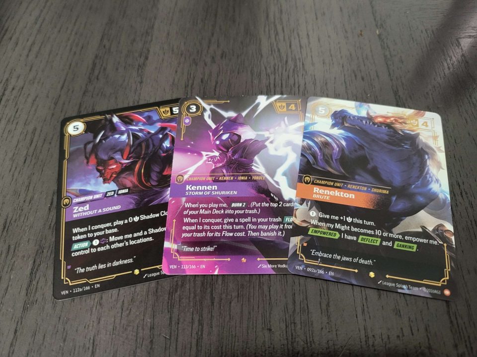 1000020573 | The Outerhaven Three K/DA-styled trading cards laid on a table: Zed on the left, Kennen in the middle, and Renekton on the right, each with colorful illustrations and stats visible at the top.