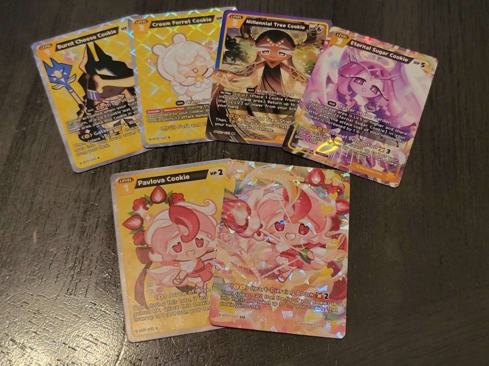 20260724_055832 | The Outerhaven Fanned collection of colorful cookie-themed trading cards on a dark table; includes Burnt Cheese Cookie, Cream Ferret Cookie, Millennial Tree Cookie, Eternal Sugar Cookie, and Pavlova Cookie cards (front two are holographic).