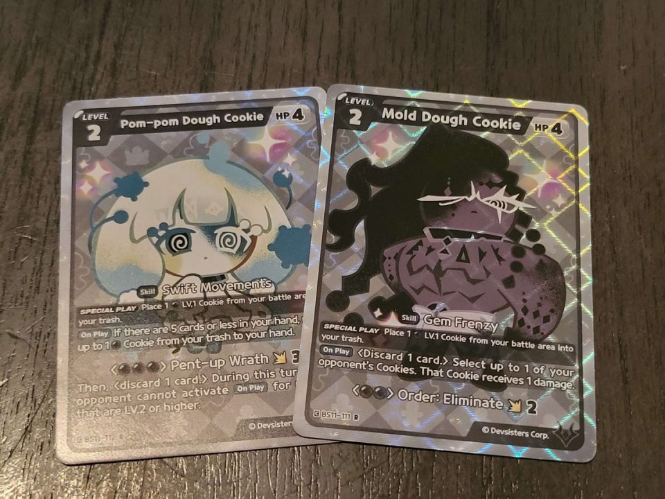 20260724_055917 | The Outerhaven Two holographic Cookie Run trading cards: Pom-pom Dough Cookie on the left and Mold Dough Cookie on the right, shown on a wooden surface.