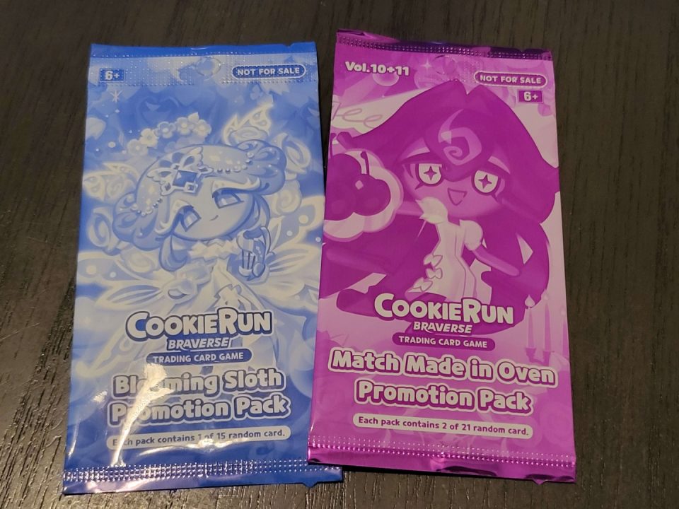 20260724_055943 | The Outerhaven Two promotional Cookie Run Braverse trading card game packs in blue and pink on a dark surface.