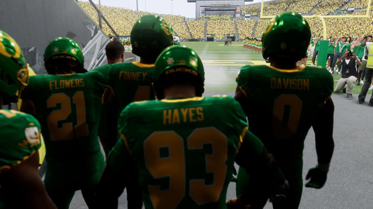 EA SPORTS™ College Football 27-Field Entry | The Outerhaven Football players in green uniforms stand on the sideline, facing the action on the field, names Flowers, Hayes, Finney, and Davison visible on jerseys behind them.