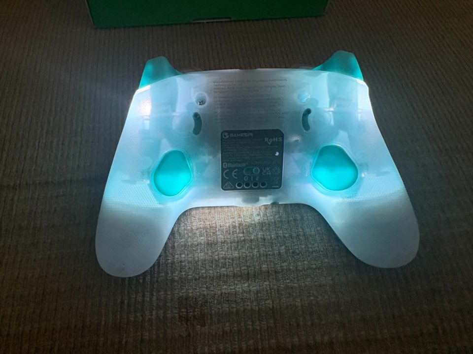 Gamesir RGB Back | The Outerhaven Back view of a translucent teal game controller with blue LED glow, resting on a carpet, showing the center label plate and grip areas.
