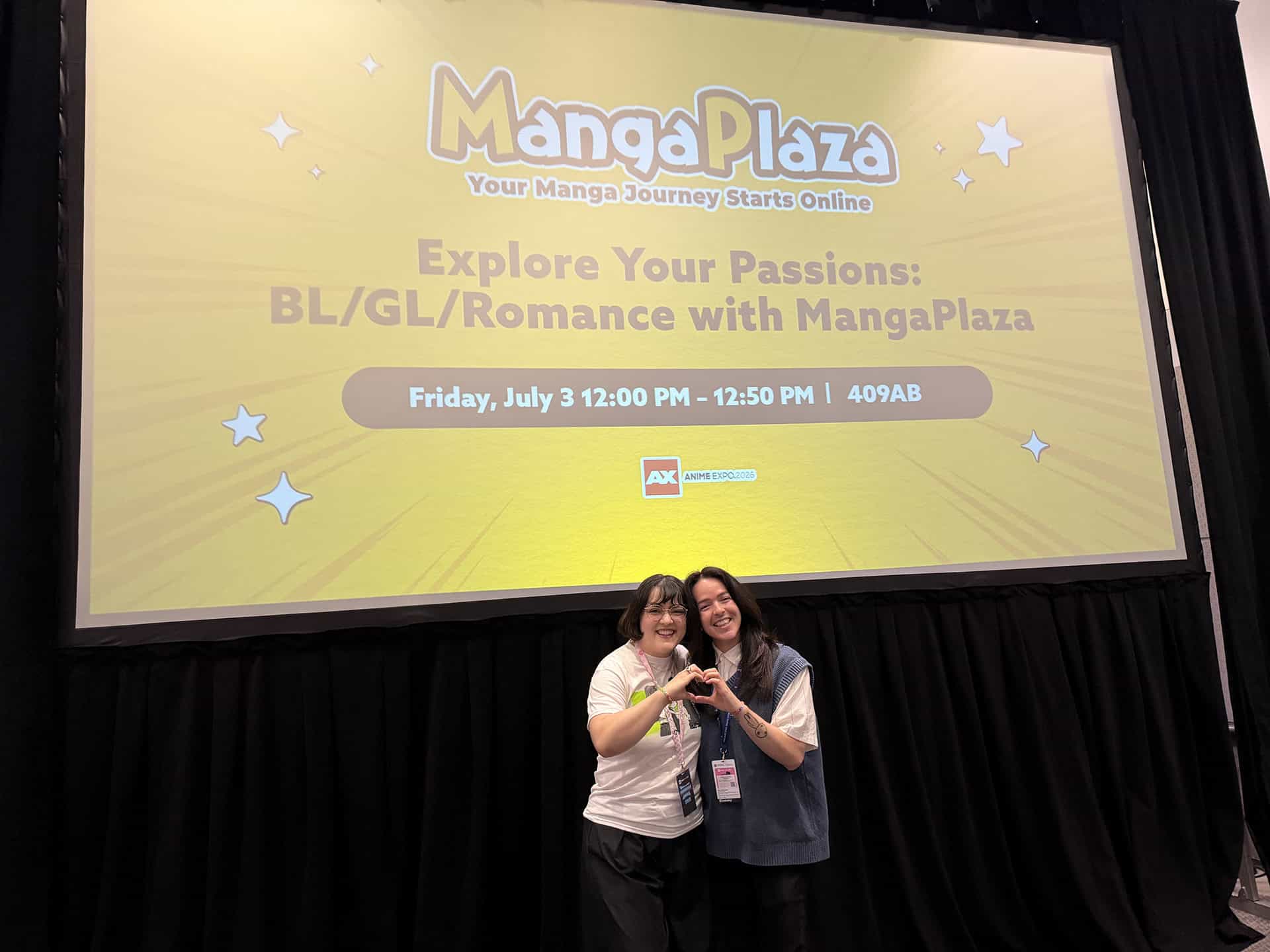 Two women smiling in front of a large yellow MangaPlaza banner, making a heart with their hands at an event stage.