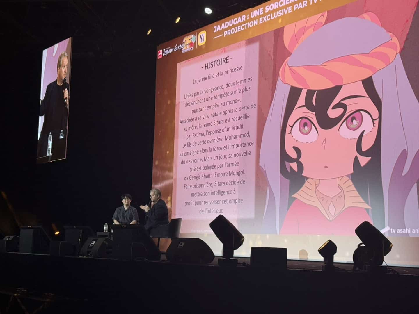 Japan Expo | The Outerhaven Two presenters sit on a dark stage beside a large projection screen showing a cartoon girl and a long French text block titled HISTOIRE.