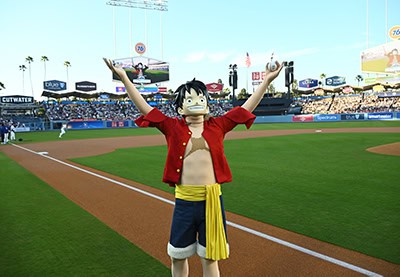One Piece | The Outerhaven Cosplayer dressed as Monkey D. Luffy raises arms on a baseball field, wearing a red open shirt and yellow sash with blue shorts, stands visible in the background.