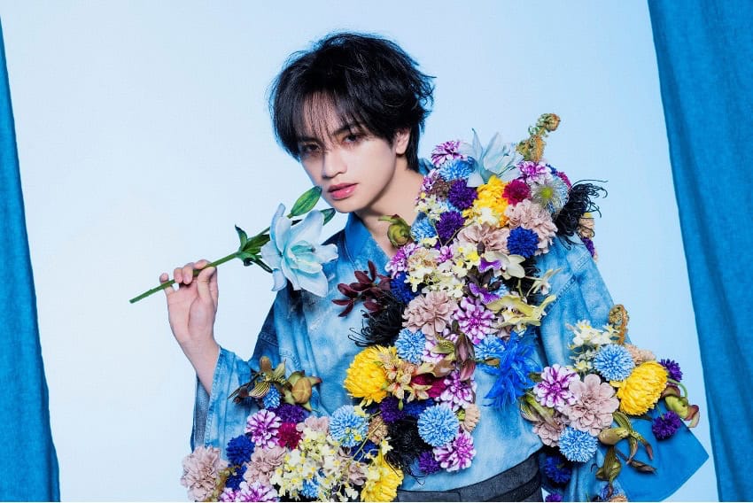 Kento Nakajima | The Outerhaven Person in a blue denim shirt poses with a large, colorful bouquet draped over their shoulder, holding a white lily against a pale blue background.