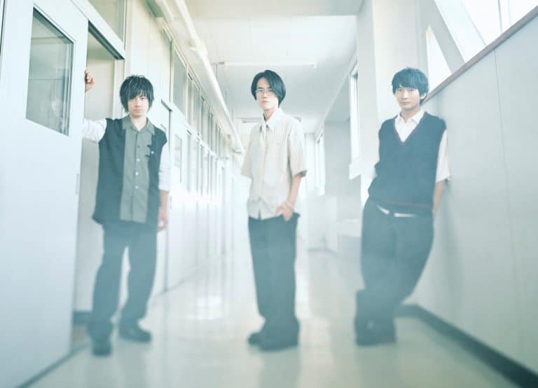 Three young men stand in a bright hallway; one leans on a door frame to the left, the others in the center and right.