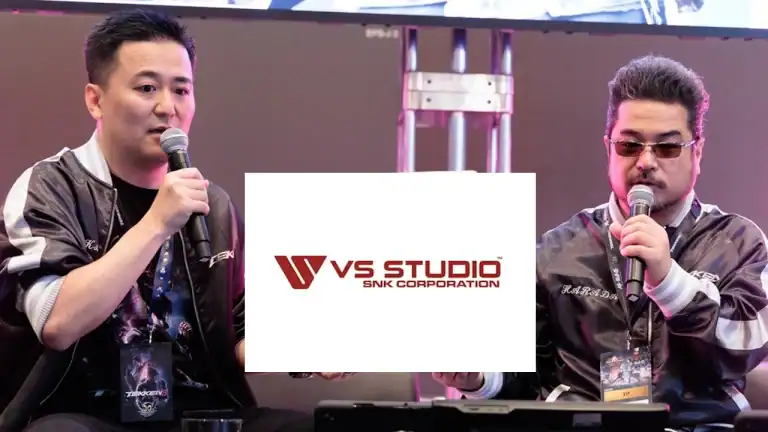 Two men on a stage speaking into microphones at a gaming event panel, with a large white VS Studio logo in the center obscuring part of the scene.
