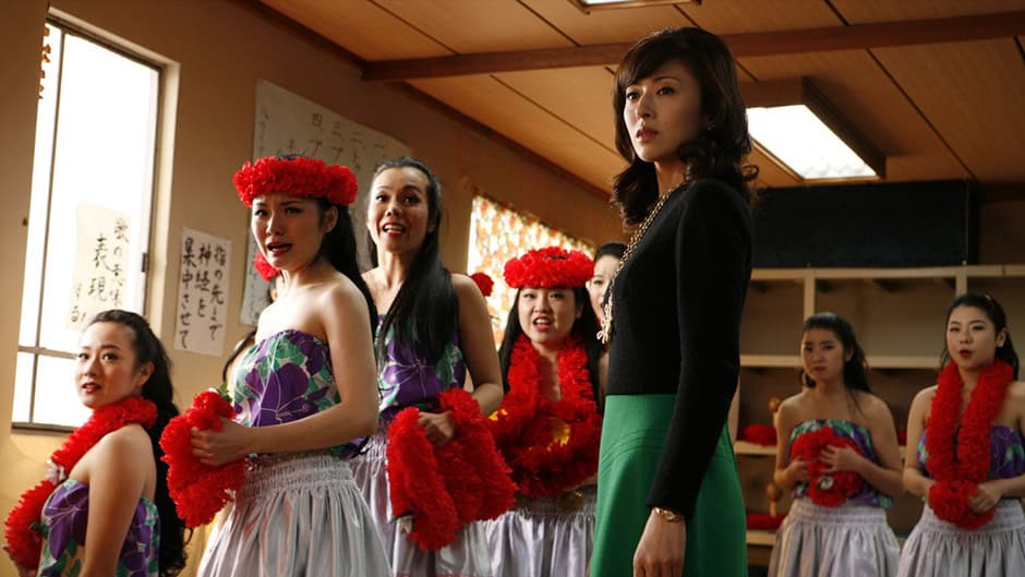 HULA GIRLS | The Outerhaven Instructor in a black top and green skirt stands at the front as a group of women in purple floral dresses and red leis practice in a dance studio.
