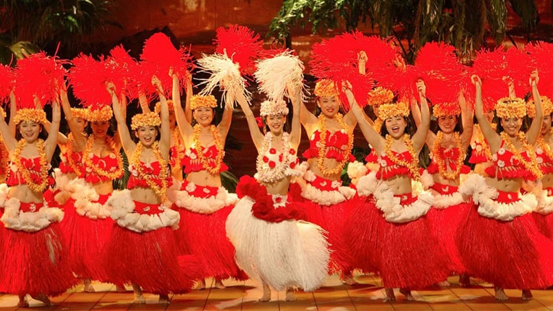 HULA GIRLS | The Outerhaven Line of female dancers in bright red and white hula skirts performing on stage with red and white feather props. Note: concise to the point about the performance.